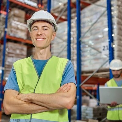 All perfectly. Young adult smiling man in protective helmet and workwear standing with folded hands looking at camera and colleague working on laptop behind