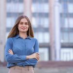 Shot of beautiful young businesswoman wearing blue chiffon shirt while standing on building background in the street with folded arms.